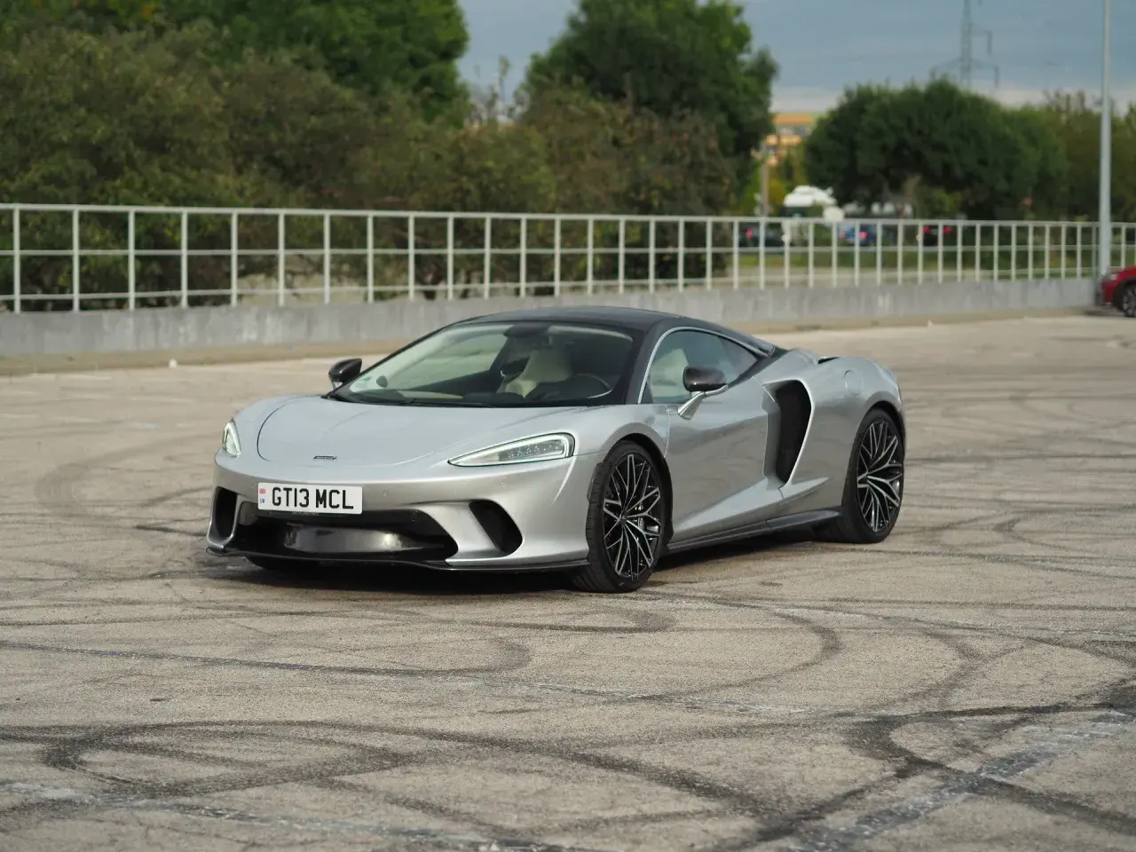 Srebrny McLaren GT z czarnym dachem, zaparkowany na placu z widocznymi śladami opon.