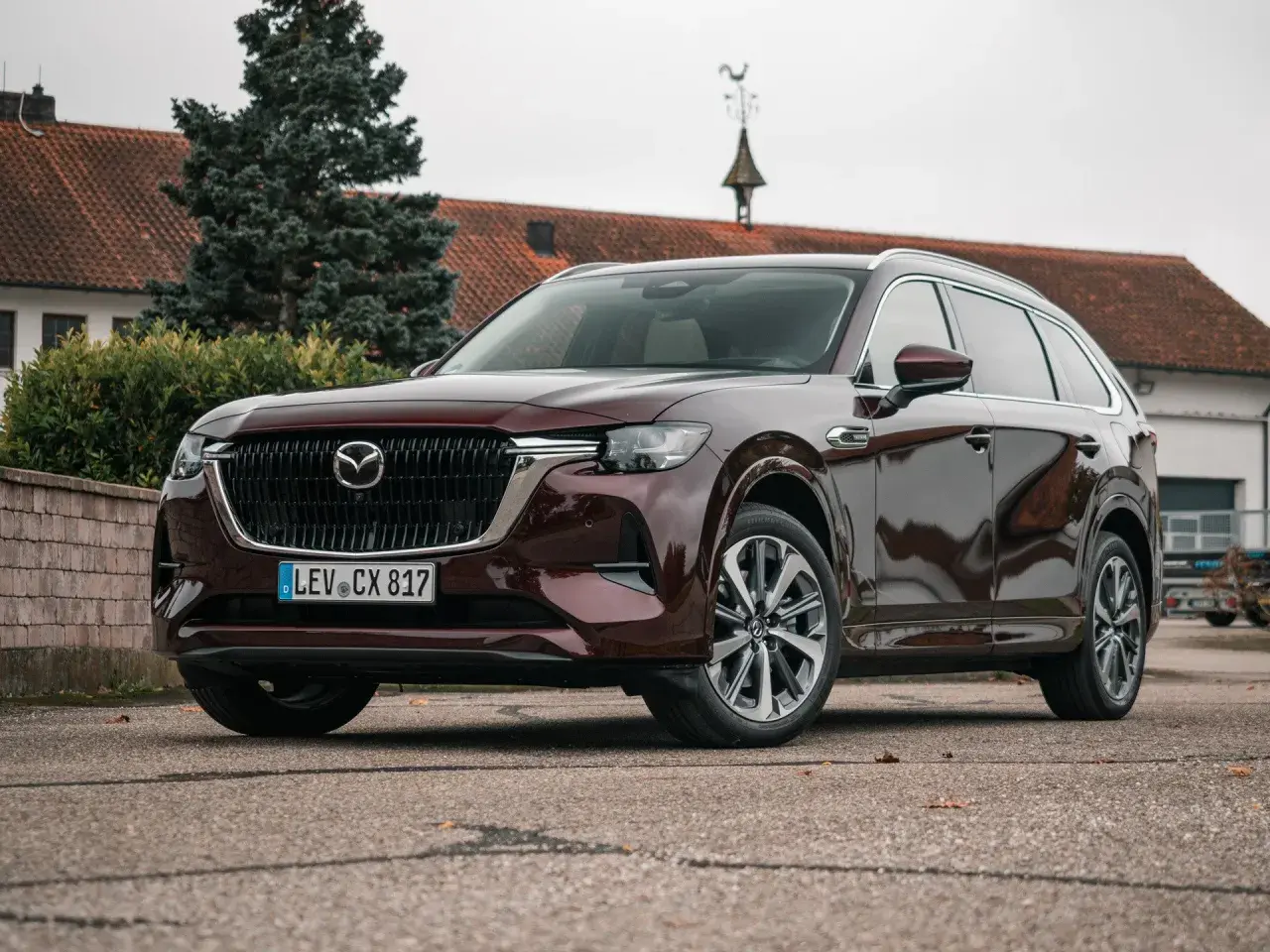Elegancka, ciemnobordowa Mazda CX-80 stoi przed budynkiem z czerwoną dachówką.