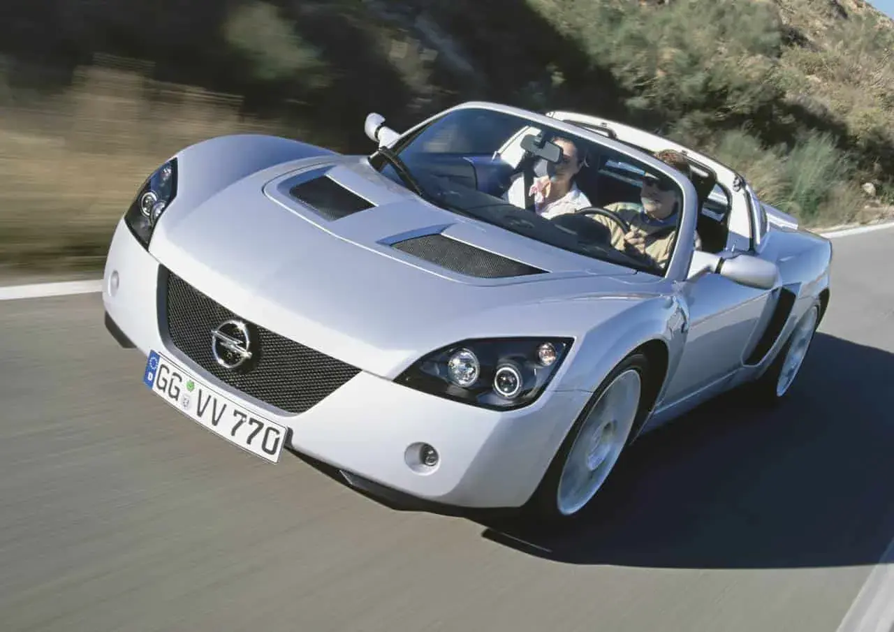 Srebrny Opel Speedster z parą na pokładzie mknie po krętej drodze, ciesząc się słońcem i wolnością.