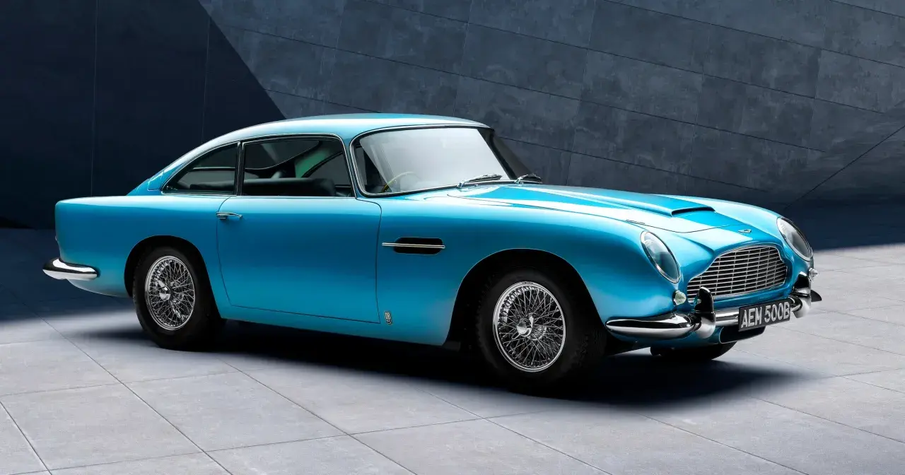 Błękitny Aston Martin DB5 z charakterystycznymi szprychowymi felgami, lśniącym chromem i elegancką linią nadwozia, zaparkowany przed nowoczesną, betonową ścianą.