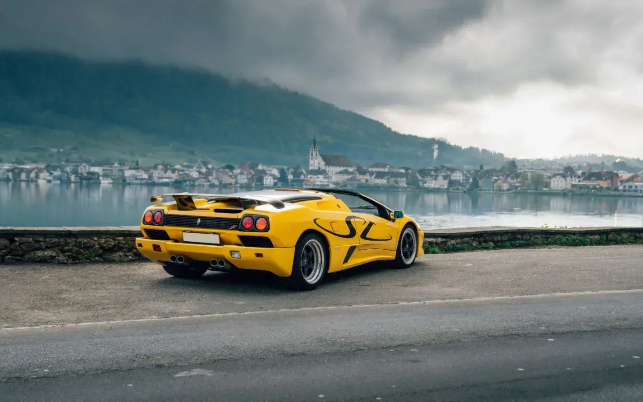 Żółty Lamborghini Diablo SV z czarnymi akcentami stoi nad jeziorem, w tle miasteczko i góry.