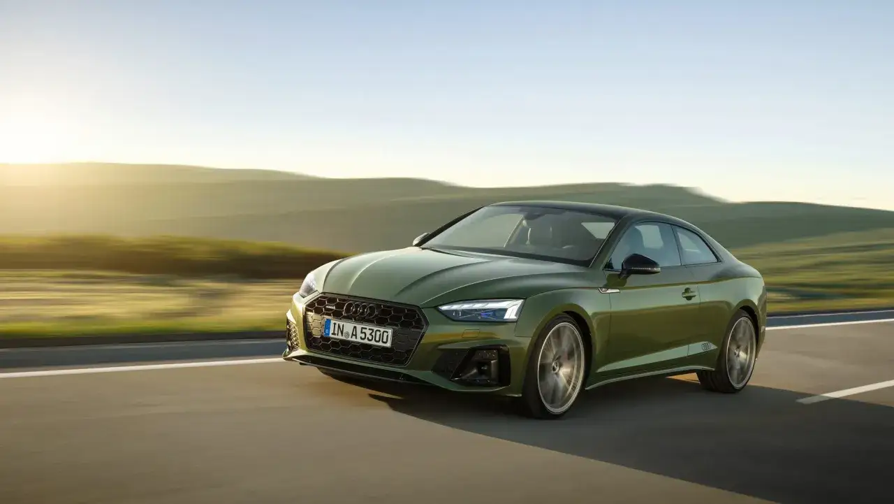 Zielony Audi A5 2013 pędzi po drodze wśród wzgórz o zachodzie słońca.