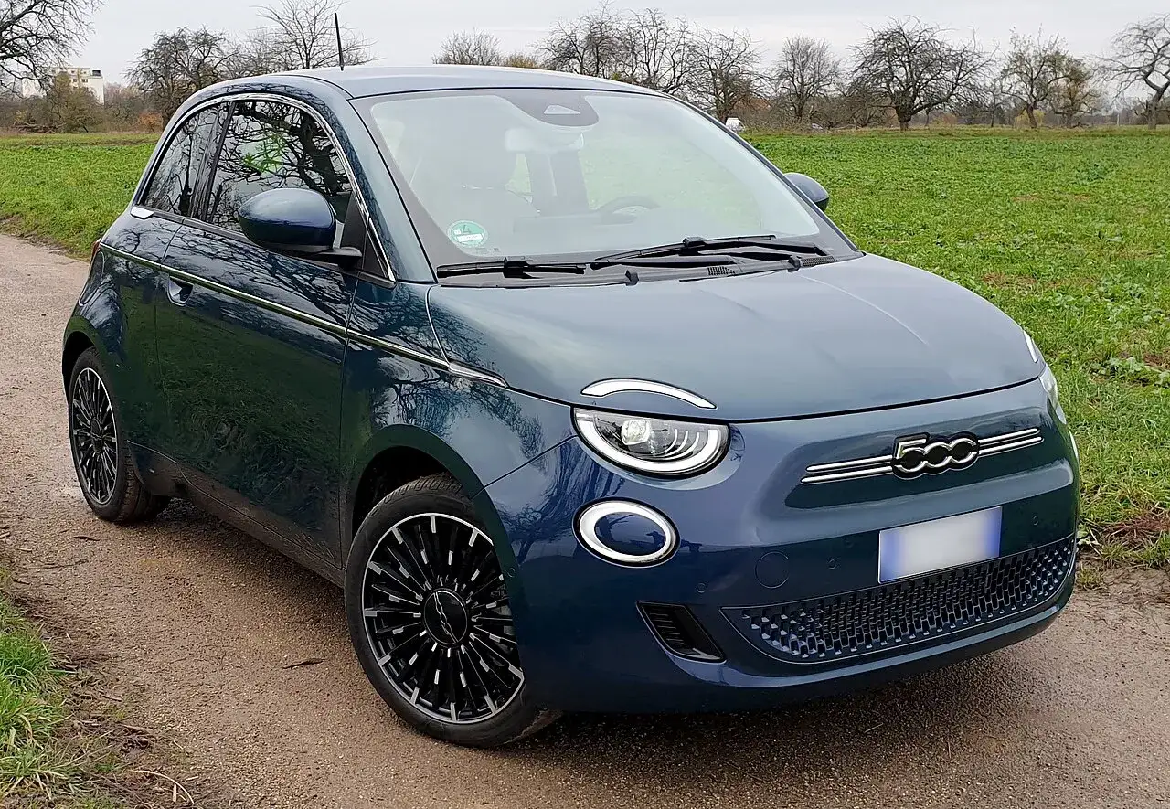 Nowy Fiat 500 w kolorze granatowym stoi na polnej drodze, otoczony zieloną trawą i drzewami.