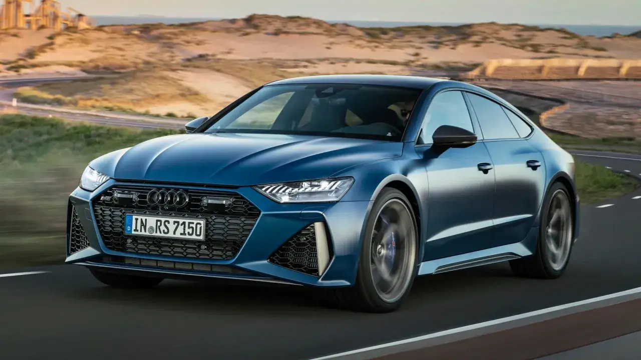 Niebieskie Audi RS7 mknie po krętej drodze, otoczone pofałdowanymi wydmami i morzem.