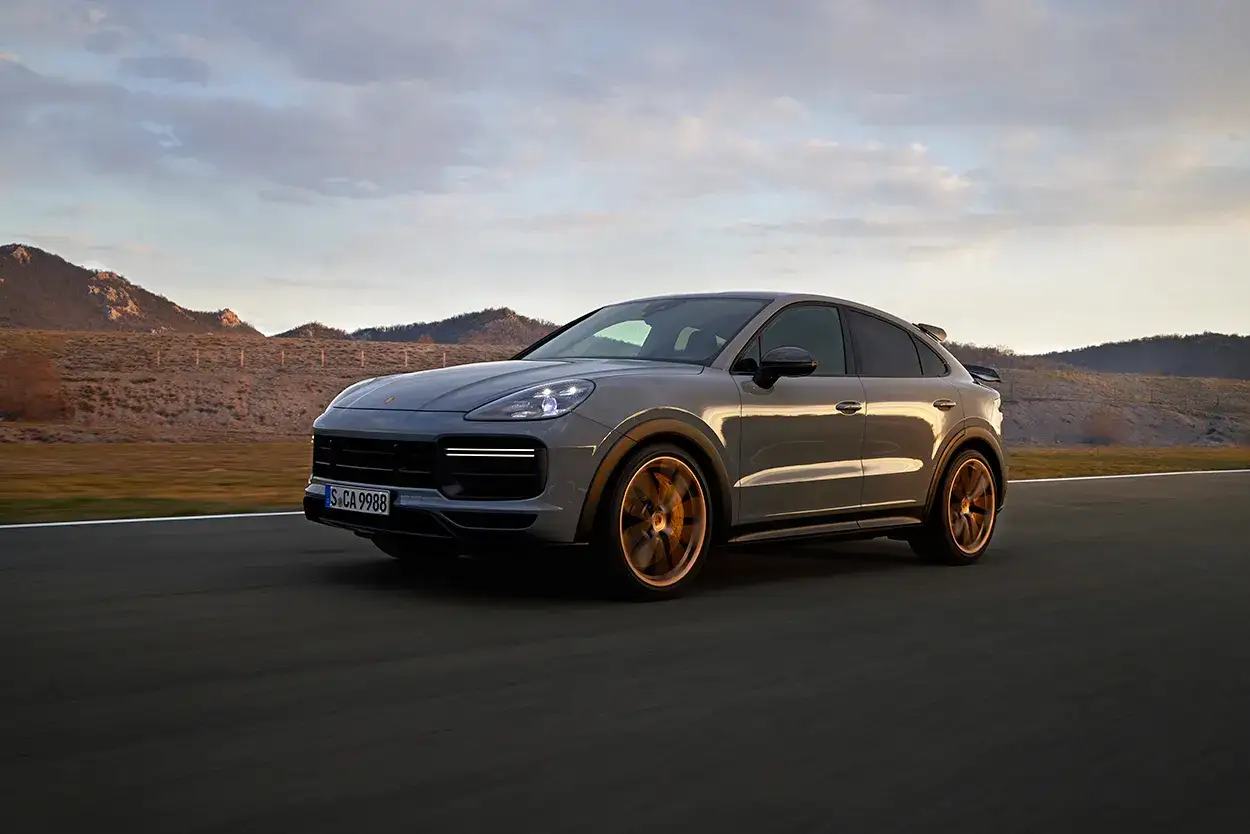 Szary Porsche Cayenne Turbo GT z brązowymi felgami pędzi po pustej drodze, otoczony surowym krajobrazem.