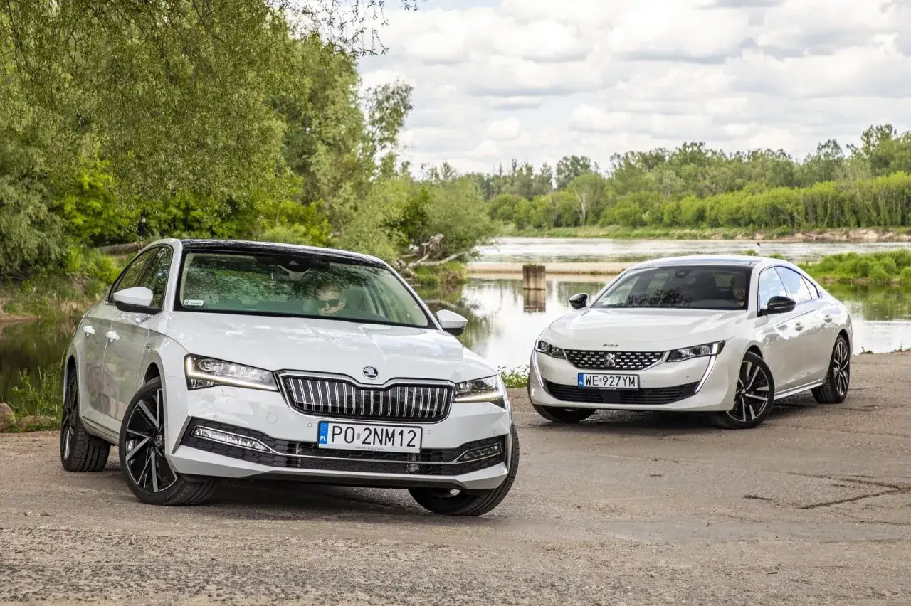 Dwa białe sedany, Skoda i Peugeot 508, stoją nad rzeką.