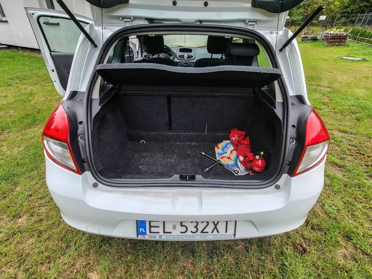 Biały Clio 3 z otwartym bagażnikiem, w którym widać gaśnicę, klucz do kół i kolorowy ręcznik.