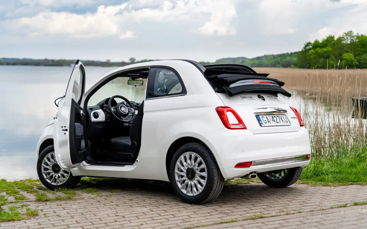 Biały Fiat 500C z otwartymi drzwiami stoi nad jeziorem, gotowy na letnią przejażdżkę.