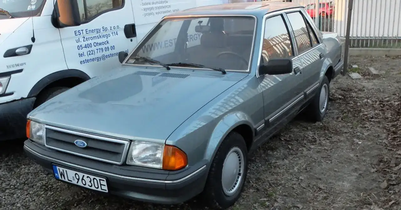 Szary Ford Orion z lat 80. stoi na zewnątrz. Widać jego przednią część, maskę i drzwi.