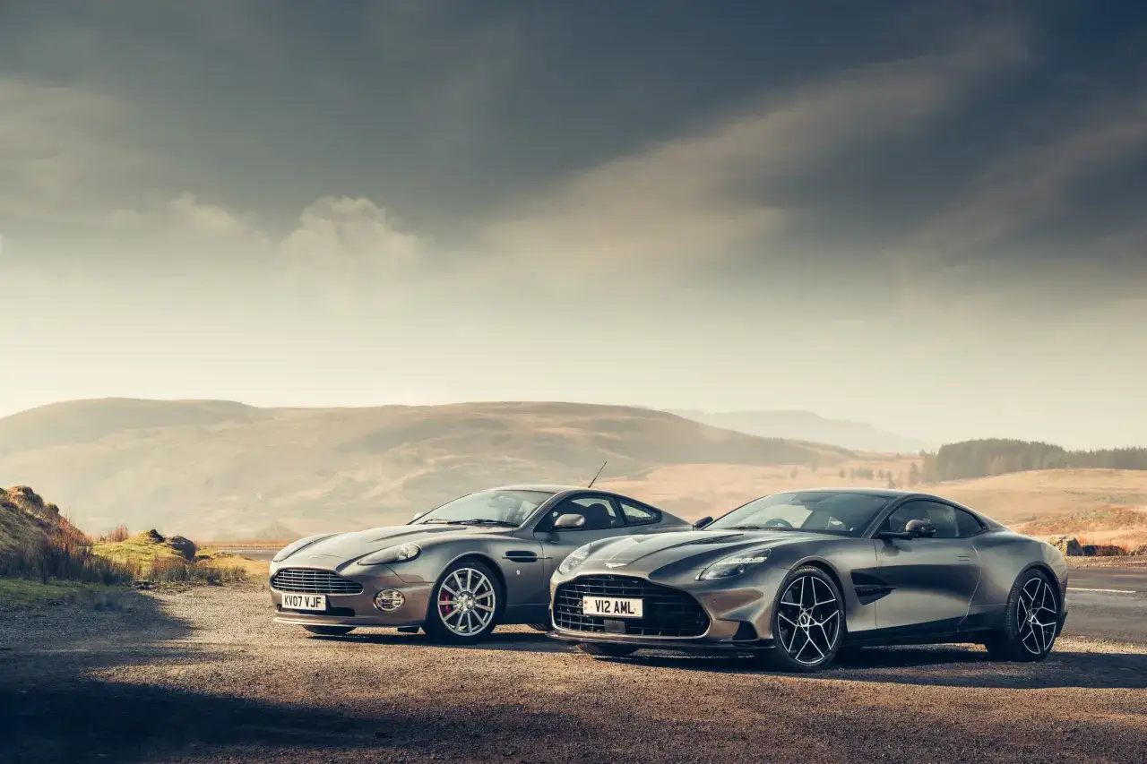 Dwa Aston Martin Vanquish, jeden starszy, drugi nowszy, stoją na tle malowniczych wzgórz.
