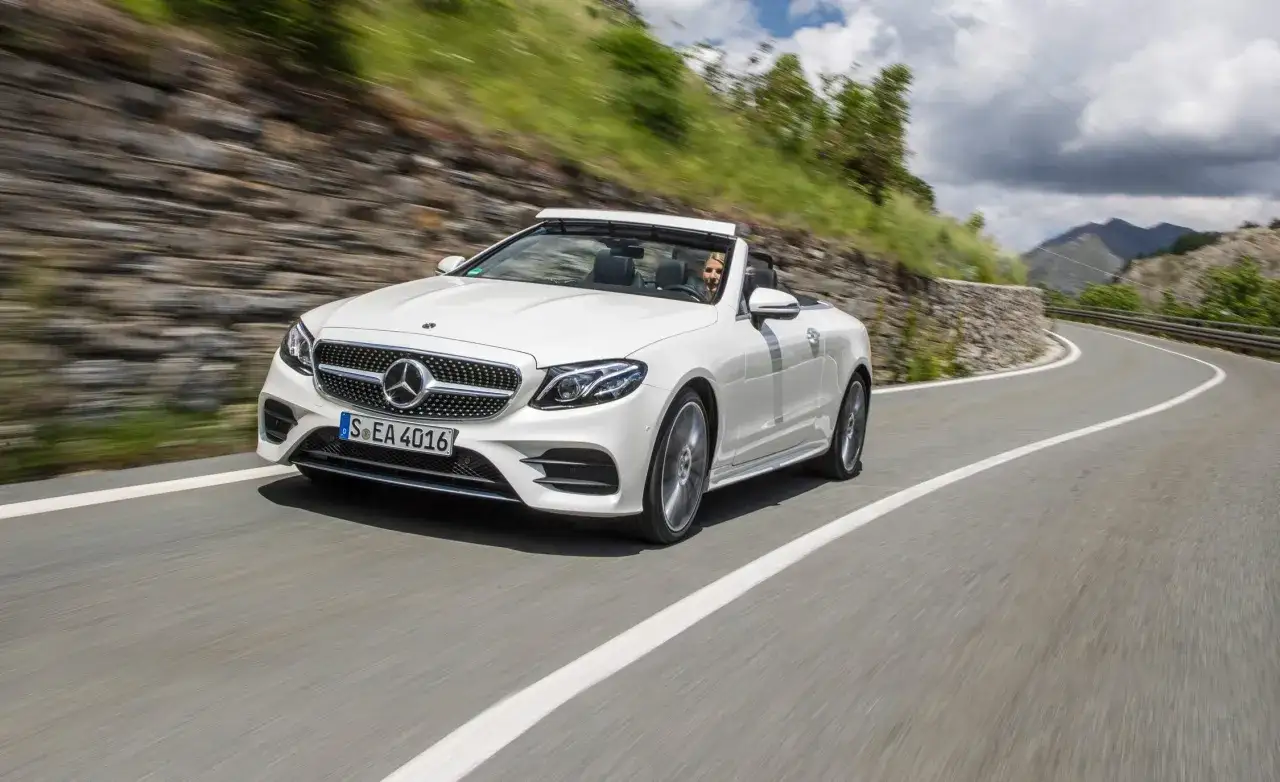 Biały kabriolet Mercedes W207 z otwartym dachem jedzie krętą górską drogą.