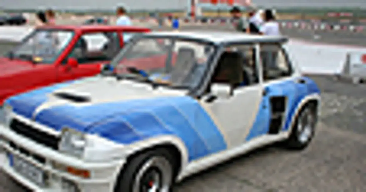 Biało-niebieski Renault 5 Turbo z charakterystycznymi naklejkami, gotowy do rajdu. Obok czerwony samochód.