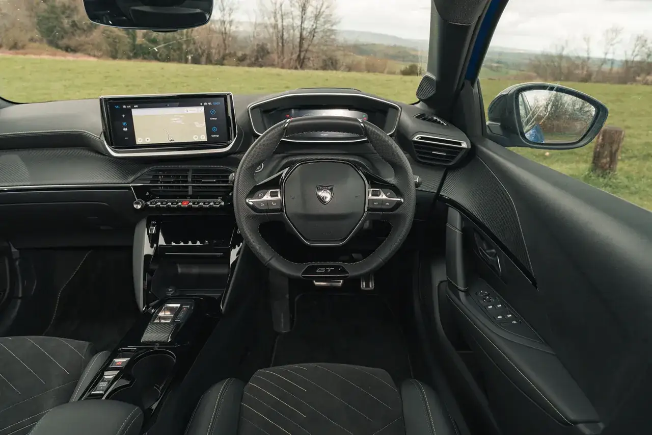 Wnętrze Peugeot e-208 z ekranem nawigacji, kierownicą GT i widokiem na zielone wzgórza.