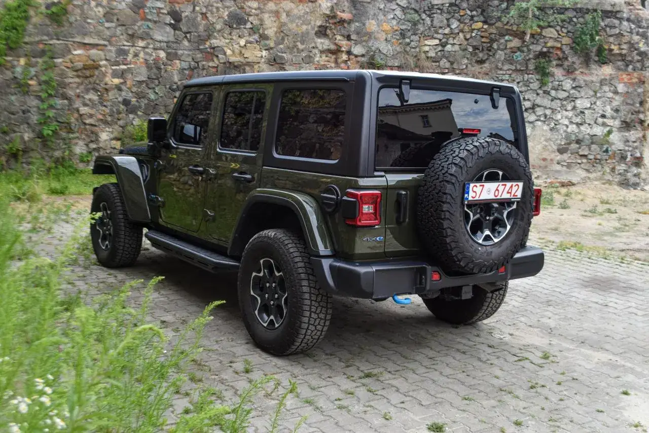 Zielony Jeep Wrangler Rubicon 4xe z zapasowym kołem z tyłu, zaparkowany przy kamiennym murze.