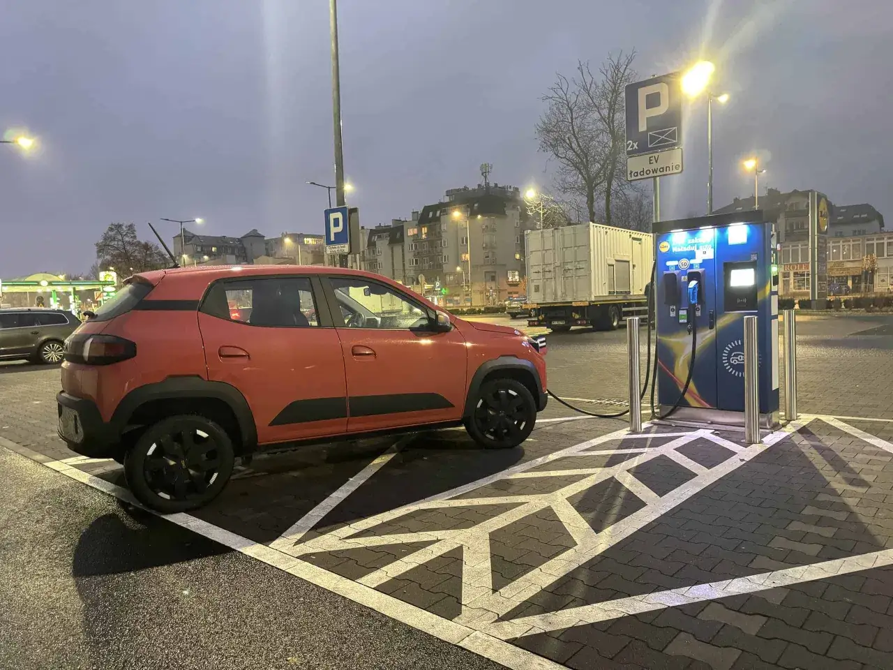Pomarańczowy Dacia Spring Electric ładuje się na stacji EV. W tle budynki i ciężarówka.