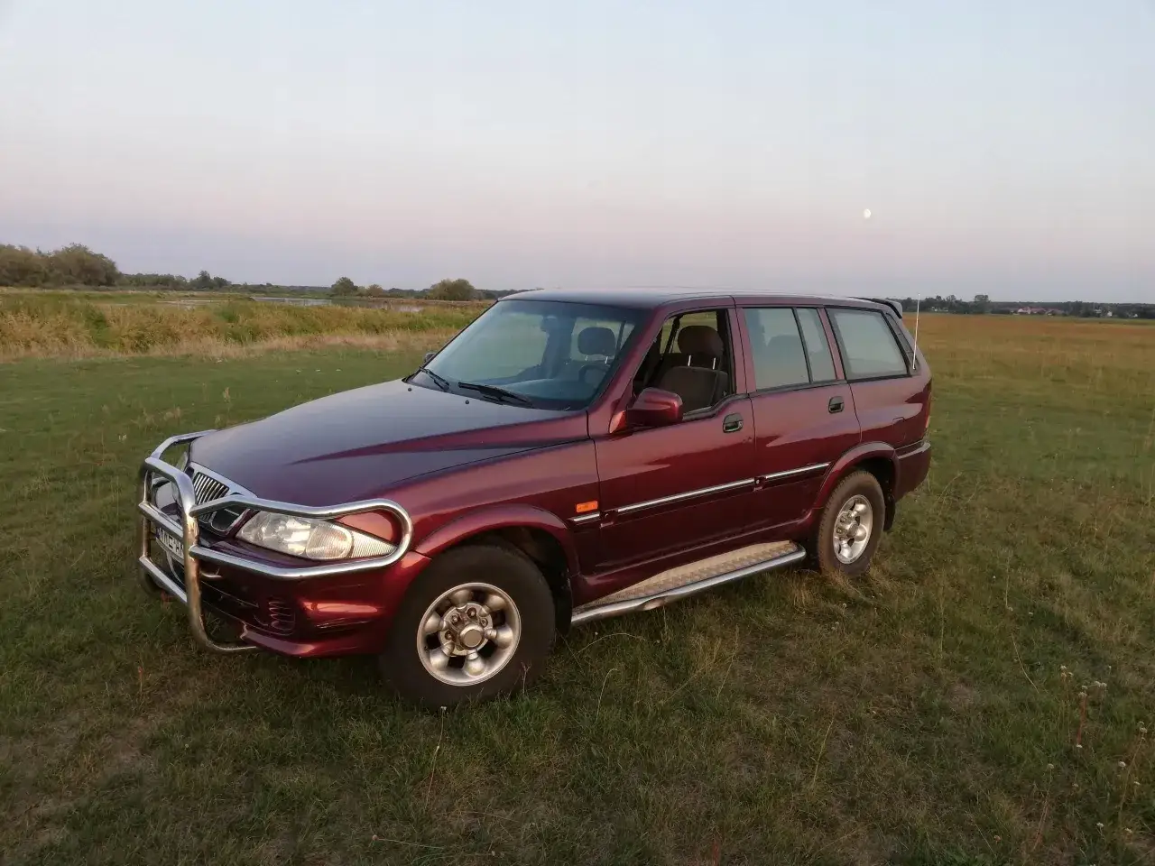Ciemnoczerwony Daewoo Musso z orurowaniem z przodu stoi na trawiastym polu. W tle jezioro i księżyc.