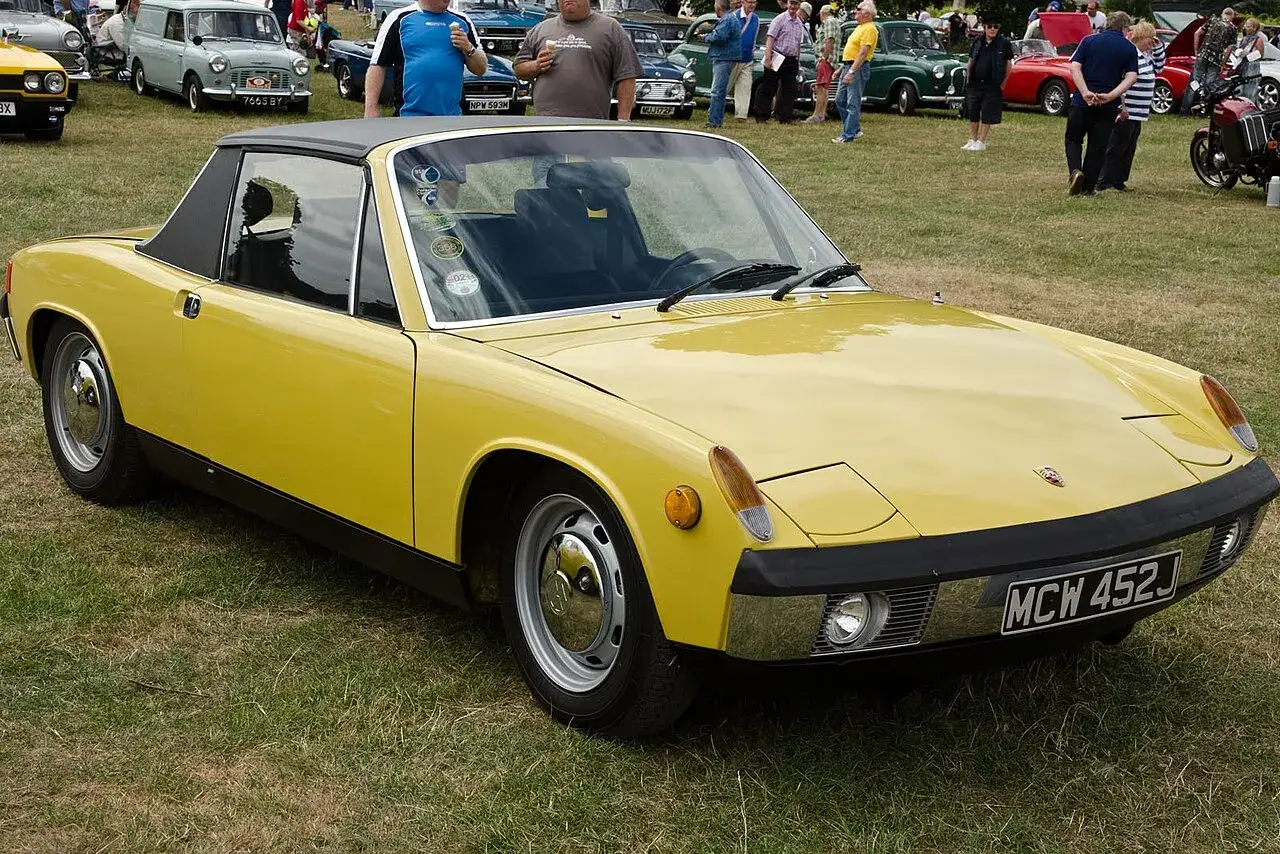 Żółty Porsche 914 z czarnym dachem stoi na trawie, obok innych klasycznych samochodów i ludzi.