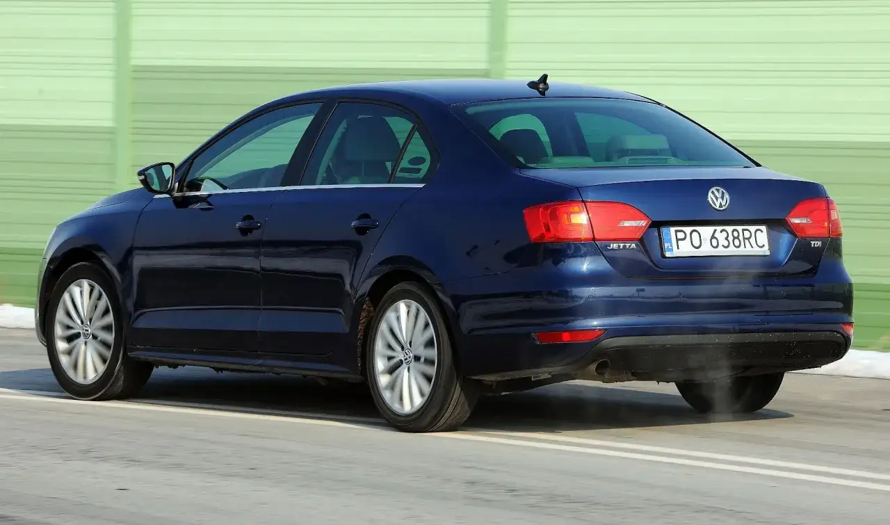 Ciemnoniebieski VW Jetta MK6 TDI z tablicą PO 638RC jedzie po drodze, z zielonym ekranem dźwiękochłonnym w tle.