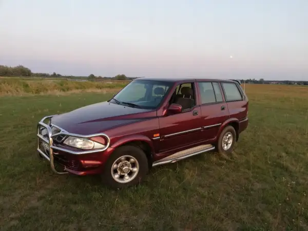 Ciemnoczerwony Daewoo Musso z orurowaniem z przodu stoi na trawiastym polu. W tle jezioro i księżyc.
