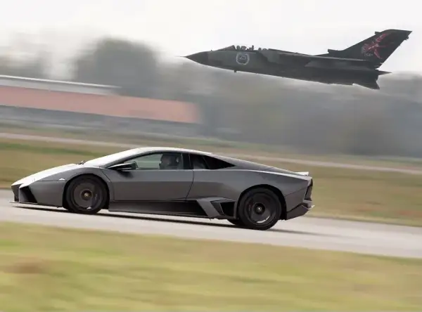 Szary Lamborghini Reventon pędzi po pasie startowym, gdy myśliwiec Tornado przelatuje nad nim.